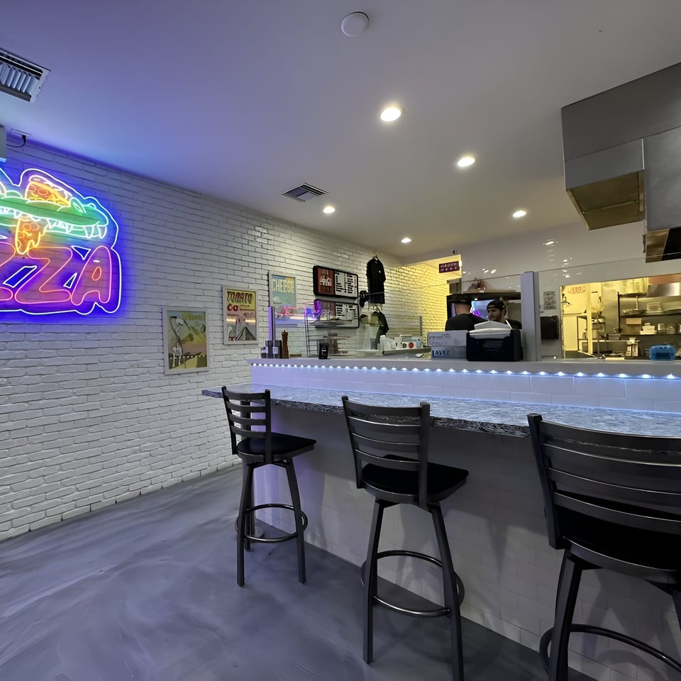 PZZA restaurant interior with a neon alligator sign, bar seating, and a kitchen in the background.