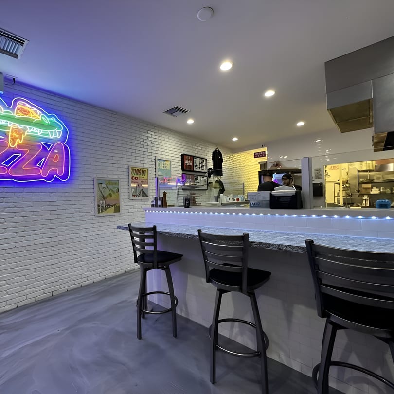 PZZA restaurant interior with a neon alligator sign, bar seating, and a kitchen in the background.