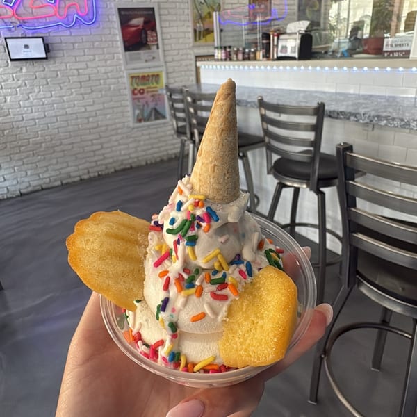 A hand holds a vanilla ice cream sundae with colorful sprinkles, a mini cone, and two madeleines.
