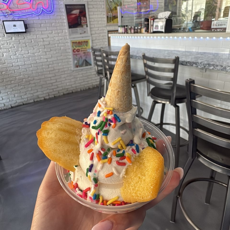 A hand holds a vanilla ice cream sundae with colorful sprinkles, a mini cone, and two madeleines.