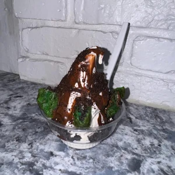 Ice cream sundae with chocolate syrup, cookie crumbles, and green cake pieces in a clear cup.