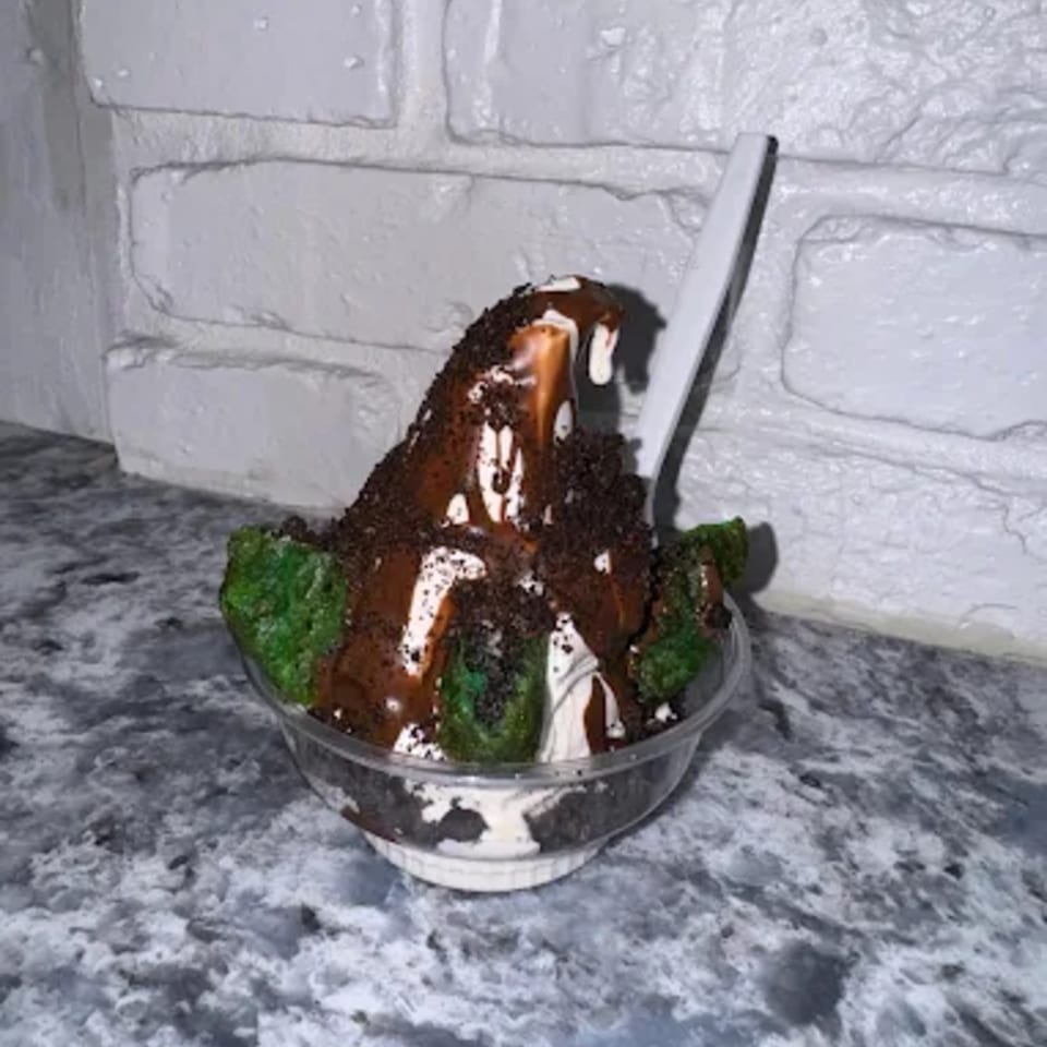 Ice cream sundae with chocolate syrup, cookie crumbles, and green cake pieces in a clear cup.