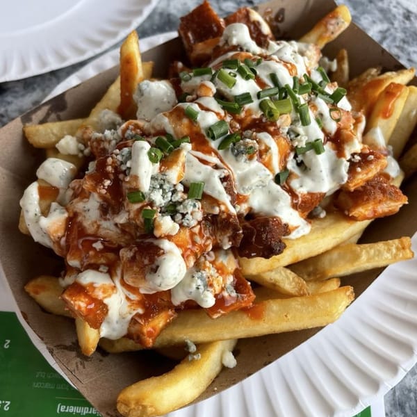 Buffalo chicken fries topped with blue cheese crumbles, ranch, and chives in a paper tray.
