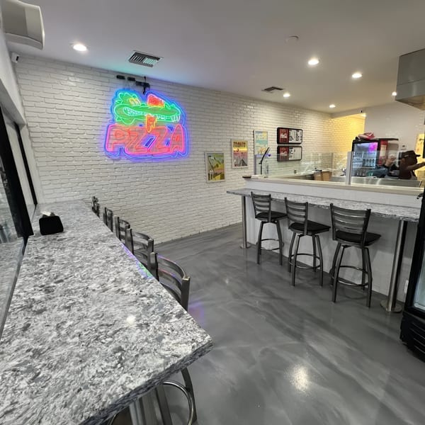 Interior of PZZA restaurant with a neon alligator sign, white brick walls, and bar seating.