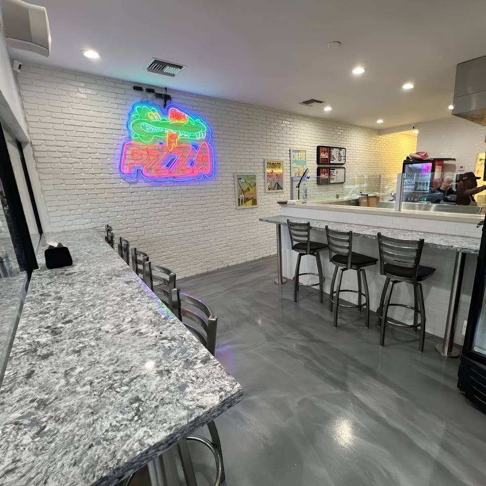 Interior of PZZA restaurant with a neon alligator sign, white brick walls, and bar seating.