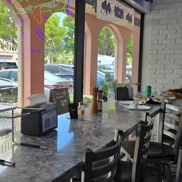 Interior view of PZZA Boca Raton, showing a counter with chairs, condiments, and a street view.