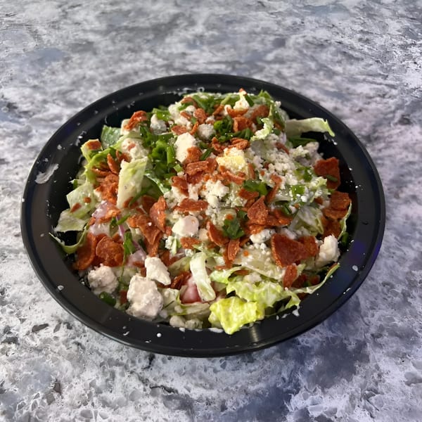 A fresh PZZA salad with lettuce, crumbled cheese, crispy pepperoni, and green onions in a black bowl.