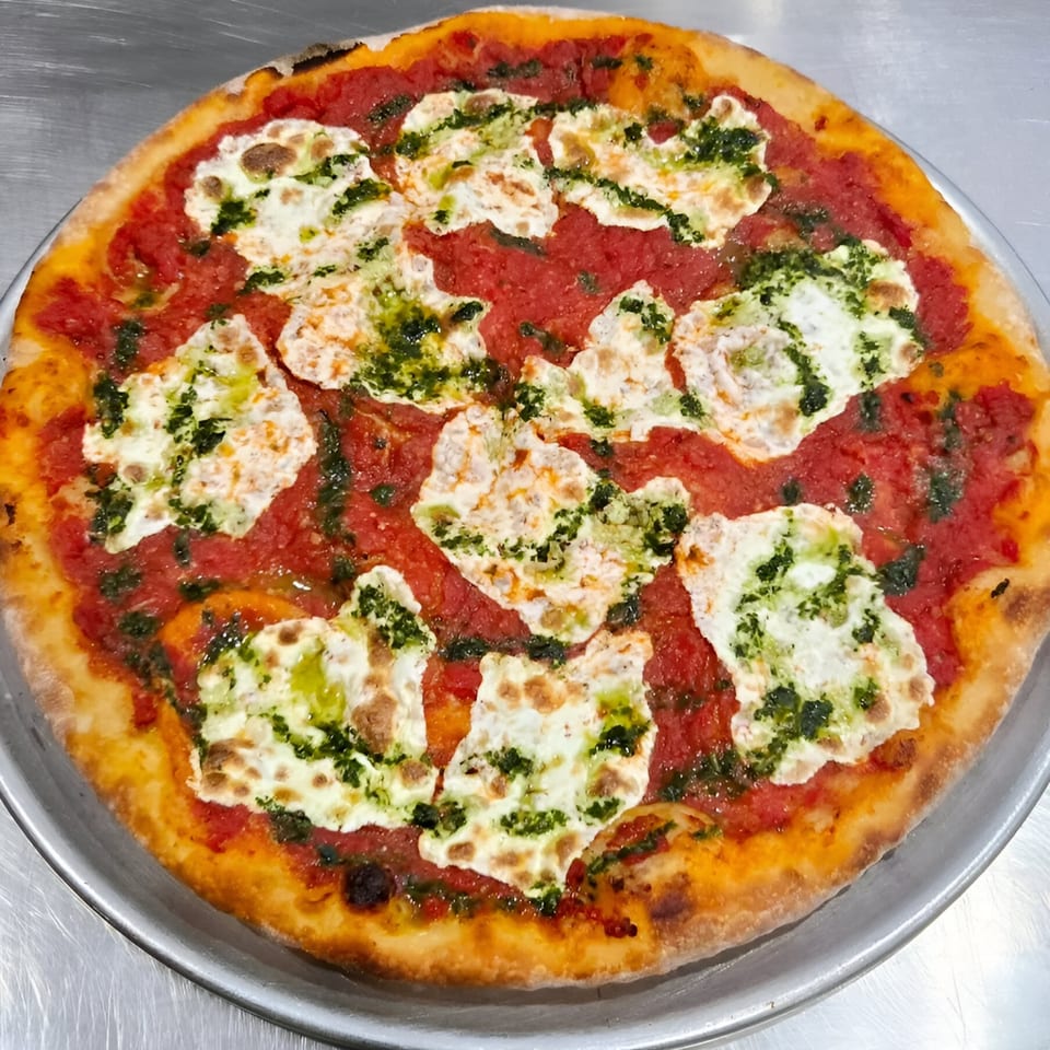 PZZA's pizza with red sauce, melted white cheese, and green pesto on a metal tray.